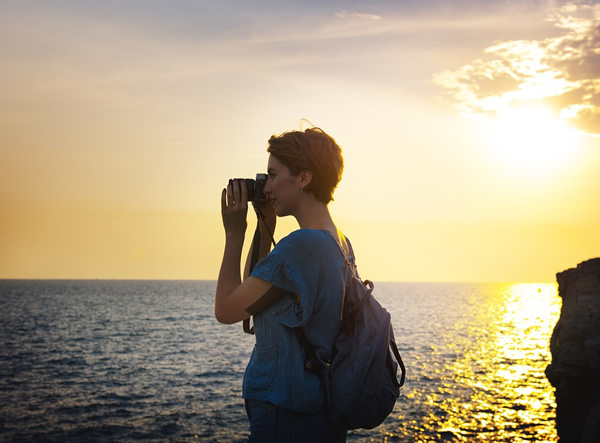 A woman traveling solo taking a photo of the ocean at sunset.