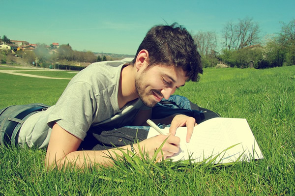 A man laying in a grass field writing in his notebook.